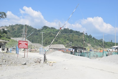 A construction or industrial site located in a mountainous area with hills in the background. A dirt road leads into the site, which is gated and marked by a sign. There are a few small buildings and green netting acting as fencing. Several people are standing near the entrance, and there are visible power lines and piles of material.