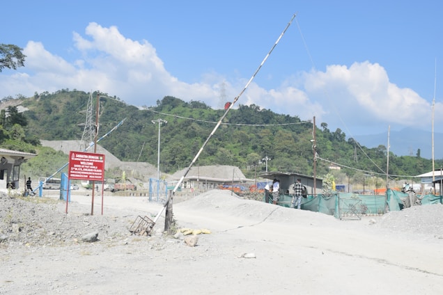 A construction or industrial site located in a mountainous area with hills in the background. A dirt road leads into the site, which is gated and marked by a sign. There are a few small buildings and green netting acting as fencing. Several people are standing near the entrance, and there are visible power lines and piles of material.
