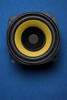 A close-up view of a speaker with a yellow diaphragm against a blue background. The speaker's circular shape is highlighted by the contrasting colors, and the image is composed to emphasize its central position.
