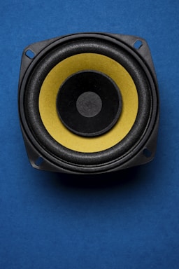A close-up view of a speaker with a yellow diaphragm against a blue background. The speaker's circular shape is highlighted by the contrasting colors, and the image is composed to emphasize its central position.