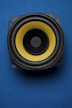 A close-up view of a speaker with a yellow diaphragm against a blue background. The speaker's circular shape is highlighted by the contrasting colors, and the image is composed to emphasize its central position.