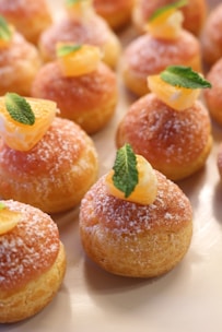 A selection of small, round pastries decorated with sugar, topped with segments of orange and fresh mint leaves. The pastries have a golden-brown color and are arranged neatly on a light-colored surface.