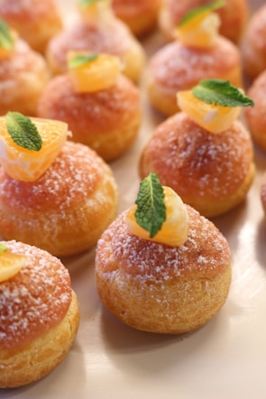 A selection of small, round pastries decorated with sugar, topped with segments of orange and fresh mint leaves. The pastries have a golden-brown color and are arranged neatly on a light-colored surface.