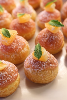 A selection of small, round pastries decorated with sugar, topped with segments of orange and fresh mint leaves. The pastries have a golden-brown color and are arranged neatly on a light-colored surface.