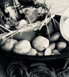 An arrangement with religious elements including a rosary and an image of Jesus in divine mercy pose. Surrounding stones are in a wooden bowl, branches form a crown-like shape, with a heart-shaped stone. A candle is placed beside the setup.