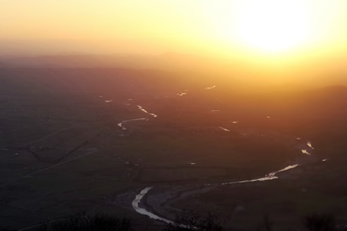 A serene landscape at sunset with a river winding through a vast, green valley. The sun is low on the horizon, casting a warm, golden glow across the scene. The mountains in the background appear shadowed and soft, adding depth to the image.