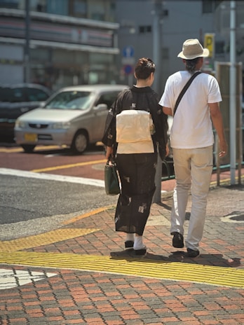A couple is walking hand in hand across a street. The woman is wearing a traditional black kimono with a white obi, while the man is dressed in a casual white shirt and hat. They are crossing at a pedestrian walkway, and several cars are visible in the background on a sunny day.