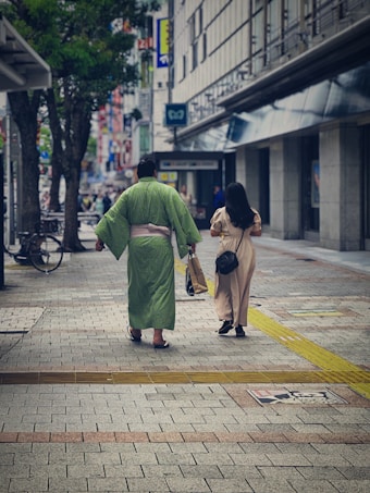 A person dressed in a traditional green kimono walks alongside another individual in casual attire on a paved street. The surroundings include modern buildings, a bicycle, and a mix of urban elements. The atmosphere is calm, featuring tree-lined pathways and signs of a bustling city scene.