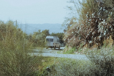 A white camper van is parked on a quiet, winding road surrounded by lush vegetation and bushes. The scene is set in a mountainous area with distant hills visible in the background, under a clear sky.