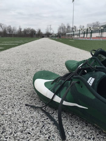 A pair of green athletic shoes with black laces lies on an artificial track field, with hurdles and bleachers visible in the background. The scene is outdoors on a cloudy day, with leafless trees surrounding the field.