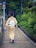 A person dressed in traditional Japanese attire walks down a paved walkway lined with lush green foliage. The path is bordered by a railing and there are traffic signs visible in the background.