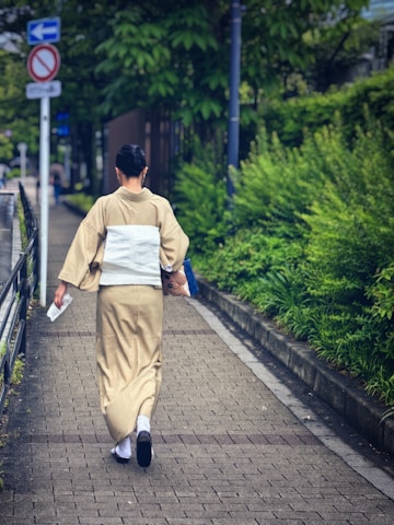 A person dressed in traditional Japanese attire walks down a paved walkway lined with lush green foliage. The path is bordered by a railing and there are traffic signs visible in the background.