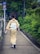 A person dressed in traditional Japanese attire walks down a paved walkway lined with lush green foliage. The path is bordered by a railing and there are traffic signs visible in the background.
