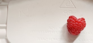 A single raspberry sits on a light-colored surface with embossed text and a recycling symbol. The berry has a vibrant red hue and is slightly heart-shaped. The surface appears to be a plastic material, possibly part of packaging.
