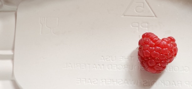A single raspberry sits on a light-colored surface with embossed text and a recycling symbol. The berry has a vibrant red hue and is slightly heart-shaped. The surface appears to be a plastic material, possibly part of packaging.