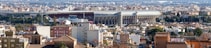 A panoramic view of a cityscape with a prominent, large stadium in the middle distance, surrounded by a mix of trees and densely packed, low-rise buildings. The stadium has a modern design and large lettering on its side. The background reveals a sprawling urban area with a variety of buildings and clear blue skies.