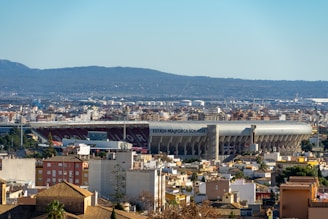 A large stadium labeled 'Estadi Mallorca Son Moix' is prominently situated in the middle of an urban landscape. The surrounding area is comprised of densely packed residential and commercial buildings. In the background, a range of hills adds depth to the scene. The sky is clear, emphasizing the sunny day.
