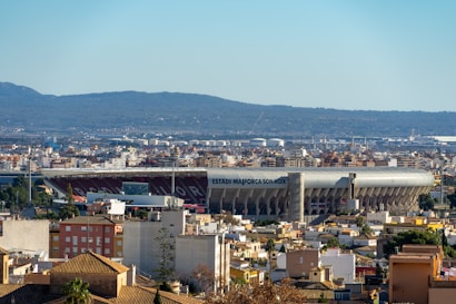 A large stadium labeled 'Estadi Mallorca Son Moix' is prominently situated in the middle of an urban landscape. The surrounding area is comprised of densely packed residential and commercial buildings. In the background, a range of hills adds depth to the scene. The sky is clear, emphasizing the sunny day.