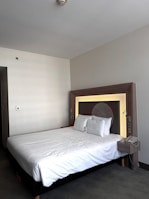 A neatly made double bed with white sheets and two pillows is placed against a modern headboard with in-built reading lights. A bedside table attached to the headboard holds a telephone. The room has a minimalist design with neutral color tones.