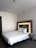 A neatly made double bed with white sheets and two pillows is placed against a modern headboard with in-built reading lights. A bedside table attached to the headboard holds a telephone. The room has a minimalist design with neutral color tones.