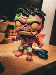 A stylized, cartoonish zombie figure with a pinkish-purple skin tone is standing on a wooden surface. The figure has exaggerated features including an open mouth with visible teeth and green patches on its body. It is dressed in tattered, grayish shorts. In the background, there are household items like a pair of scissors and some baskets.
