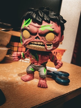 A stylized, cartoonish zombie figure with a pinkish-purple skin tone is standing on a wooden surface. The figure has exaggerated features including an open mouth with visible teeth and green patches on its body. It is dressed in tattered, grayish shorts. In the background, there are household items like a pair of scissors and some baskets.