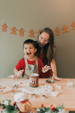 A child and an adult, both dressed in red clothing, are happily engaged in a baking activity. The child is wearing an apron and has flour on their hands, while a festive tin is placed on a flour-covered wooden table. In the background, a garland of gingerbread man decorations hangs on a light-colored wall.