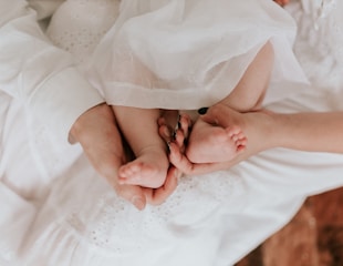 A tender moment captured with an adult gently cradling a baby's feet in their hands. The soft, white fabric creates a serene atmosphere, highlighting the contrast between the adult's and baby's skin.