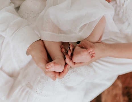 A tender moment captured with an adult gently cradling a baby's feet in their hands. The soft, white fabric creates a serene atmosphere, highlighting the contrast between the adult's and baby's skin.