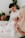 A tender family moment with a mother and father holding a baby dressed in white. The setting appears cozy, with soft natural light and indoor plants in the background. The mother and father look down lovingly at the baby.
