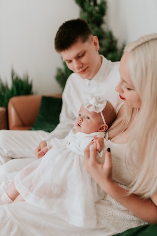 A tender family moment with a mother and father holding a baby dressed in white. The setting appears cozy, with soft natural light and indoor plants in the background. The mother and father look down lovingly at the baby.