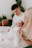 A tender family moment with a mother and father holding a baby dressed in white. The setting appears cozy, with soft natural light and indoor plants in the background. The mother and father look down lovingly at the baby.