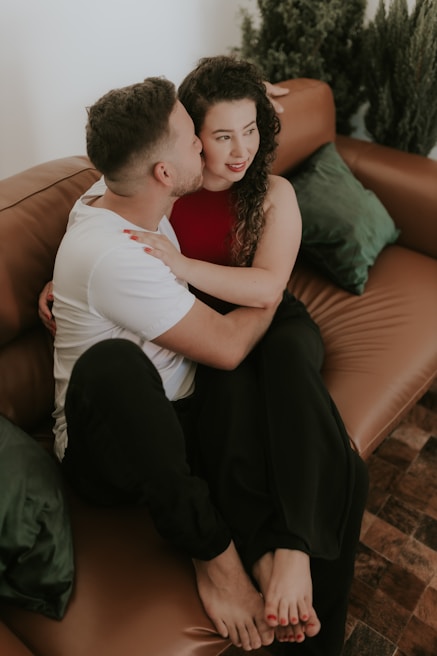 A couple is sitting closely on a brown leather sofa. The woman, wearing a red top and black pants, has dark curly hair and red lipstick. The man, wearing a white t-shirt and black pants, is embracing her and appears to be whispering or kissing her cheek. Green cushions and potted plants add to the cozy setting.
