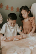 Two children are engaged in a baking activity, rolling dough on a wooden table. Flour is scattered around, and there are gingerbread decorations in the background.