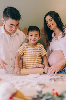 A family in a kitchen setting is engaged in baking, with a child happily using a rolling pin to flatten dough. The parents, one wearing a white shirt and the other a white top with a visible pregnant belly, are assisting and smiling.