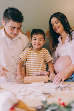A family in a kitchen setting is engaged in baking, with a child happily using a rolling pin to flatten dough. The parents, one wearing a white shirt and the other a white top with a visible pregnant belly, are assisting and smiling.