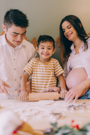 A family in a kitchen setting is engaged in baking, with a child happily using a rolling pin to flatten dough. The parents, one wearing a white shirt and the other a white top with a visible pregnant belly, are assisting and smiling.