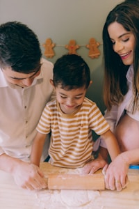 A happy family scene with a child and two adults engaging in baking activities together. The child is in the center, smiling while using a rolling pin to flatten dough on a table covered with flour. The adults, one on either side, are guiding and assisting the child with the baking process. Gingerbread man decorations hang in the background, adding a festive touch.