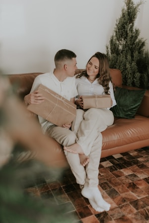 A couple is sitting on a brown leather couch in a cozy living room. Both individuals are holding wrapped presents and appear to be smiling at each other. A green pillow is beside them, and there is a Christmas tree in the background. The scene is framed with a warm and festive ambiance.