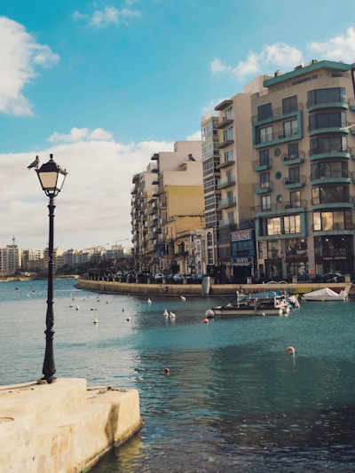 A seaside urban scene with a row of modern apartment buildings lining the waterfront. A lamp post with birds perched on it stands in the foreground, overlooking a calm body of water with several boats and buoys. The sky is clear with a few clouds, and the overall setting conveys a sense of tranquility.