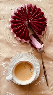 A beautifully designed cake with a deep red, glossy surface featuring star-like pleats sits on a transparent plate. A slice has been removed, exposing its layered interior of white, pink, and brown hues. Beside the cake, a cup of coffee rests on a matching saucer, with a fork lying next to the cake slice. The background is adorned with a subtle, floral-patterned tablecloth.