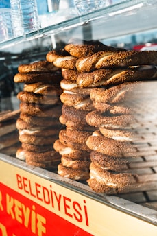 Stacks of Turkish simit, a ring-shaped bread coated with sesame seeds, are displayed inside a glass case. Several plastic water bottles are placed on top of the case. The sign 'Belediyesi' in red lettering is visible at the bottom.