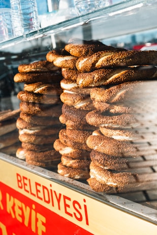 Stacks of Turkish simit, a ring-shaped bread coated with sesame seeds, are displayed inside a glass case. Several plastic water bottles are placed on top of the case. The sign 'Belediyesi' in red lettering is visible at the bottom.