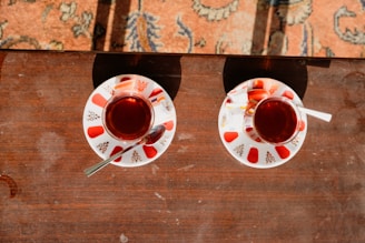 Two small glass cups filled with dark tea placed on a wooden table. Each cup is on a decorative saucer embellished with red patterns and accompanied by a metal spoon and a sugar packet. Sunlight casts shadows on the table and part of a patterned carpet is visible in the background.