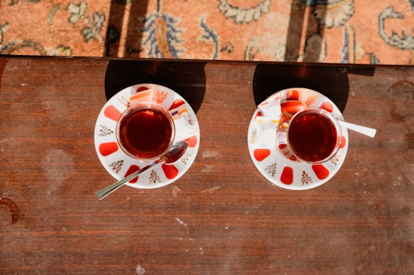 Two small glass cups filled with dark tea placed on a wooden table. Each cup is on a decorative saucer embellished with red patterns and accompanied by a metal spoon and a sugar packet. Sunlight casts shadows on the table and part of a patterned carpet is visible in the background.