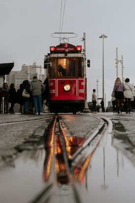 A vintage red tram with the sign 'Taksim Tünel' prominently displayed is moving along wet tram tracks, with its reflection visible in a puddle. The surrounding area is bustling with people, some waiting for the tram to pass or board. Various buildings and street lights are in the background, under an overcast sky.