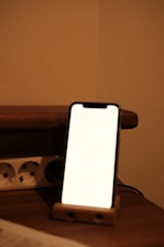 A smartphone with a blank white screen is placed on a wooden stand on a table. There is a power strip with sockets in the background against a beige wall, creating a softly lit ambiance.