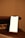 A smartphone with a blank white screen is placed on a wooden stand on a table. There is a power strip with sockets in the background against a beige wall, creating a softly lit ambiance.