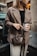 A person stands near an old vehicle, holding a brown textured handbag and a take-away coffee cup labeled 'QUADOR'. The person is dressed in a stylish brown blazer over a black top and light-colored pants.