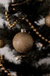 A close-up view of an artificial Christmas tree adorned with a sparkling gold ornament and a string of golden beads. The tree has dark green branches that fill the background.
